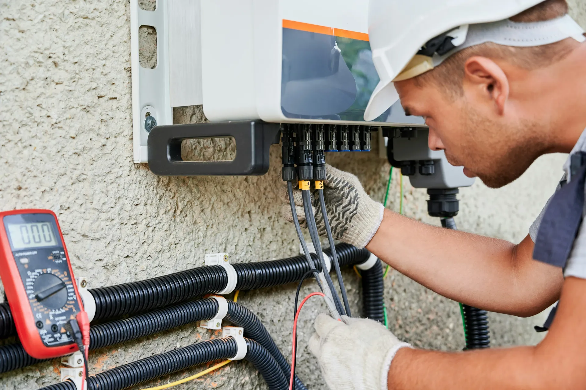 An electrician working on a solar inverter, connecting cables and checking readings with a red multimeter on a textured wall.