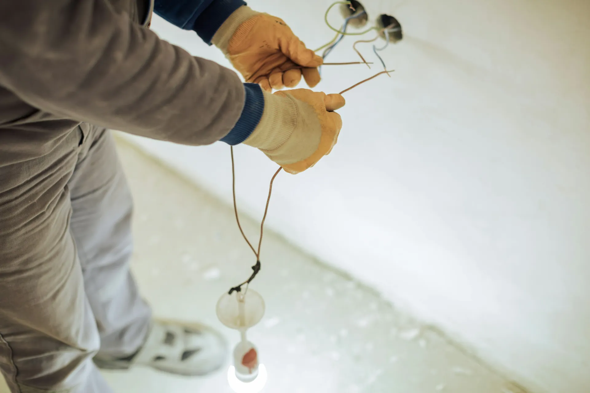 Close-up of an electrician's hands in yellow gloves, installing new electrical wiring for a light fixture in a wall or ceiling during construction, with a temporary bulb illuminating the work area.