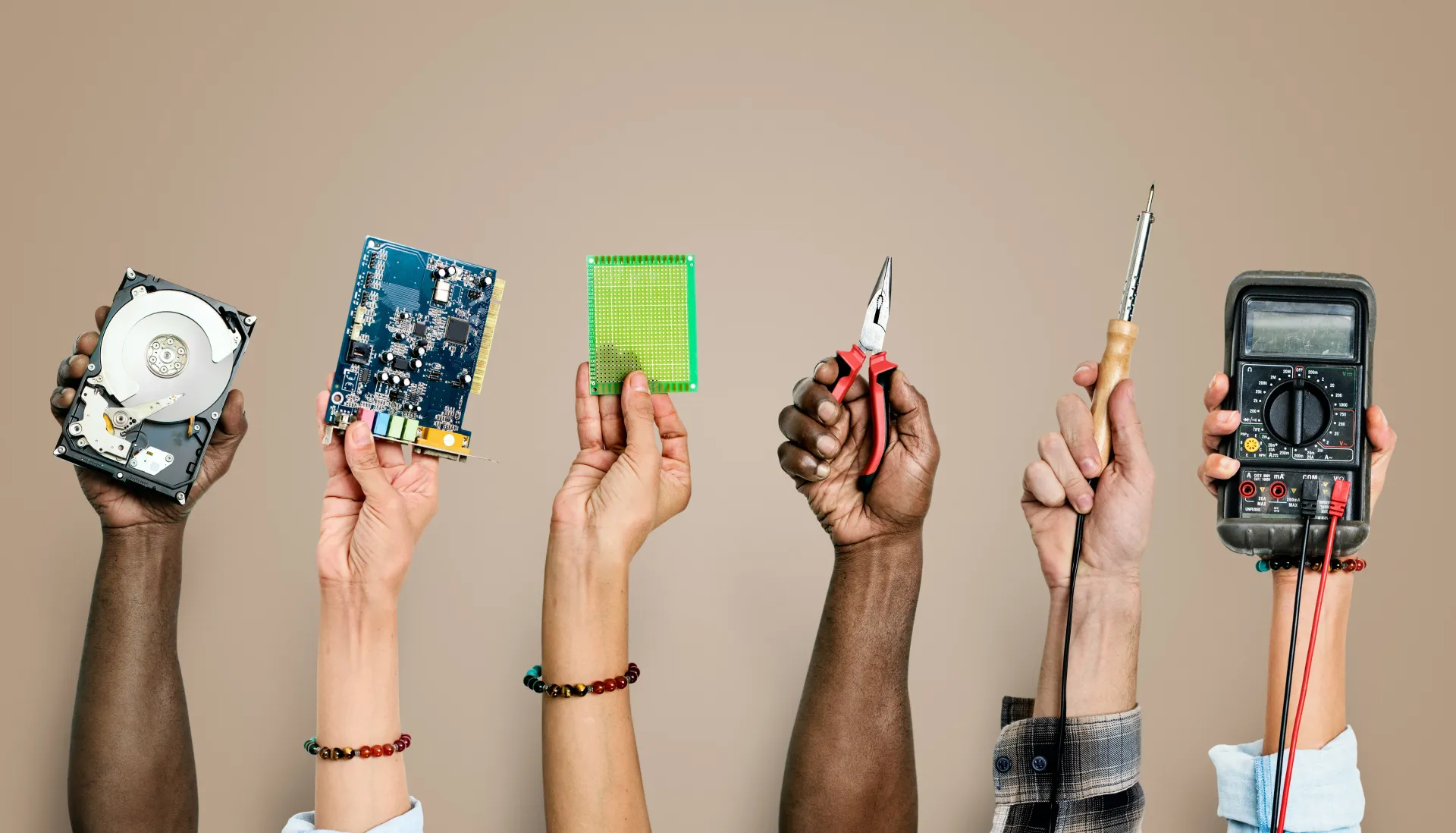 Hands holding various electrical and electronic tools and components such as a hard drive, circuit boards, pliers, a soldering iron, and a multimeter.